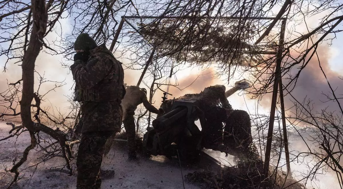 soldați, îmbrăcați în uniforme de camuflaj, operează un tun de artilerie ascuns sub o plasă de camuflaj printre crengi