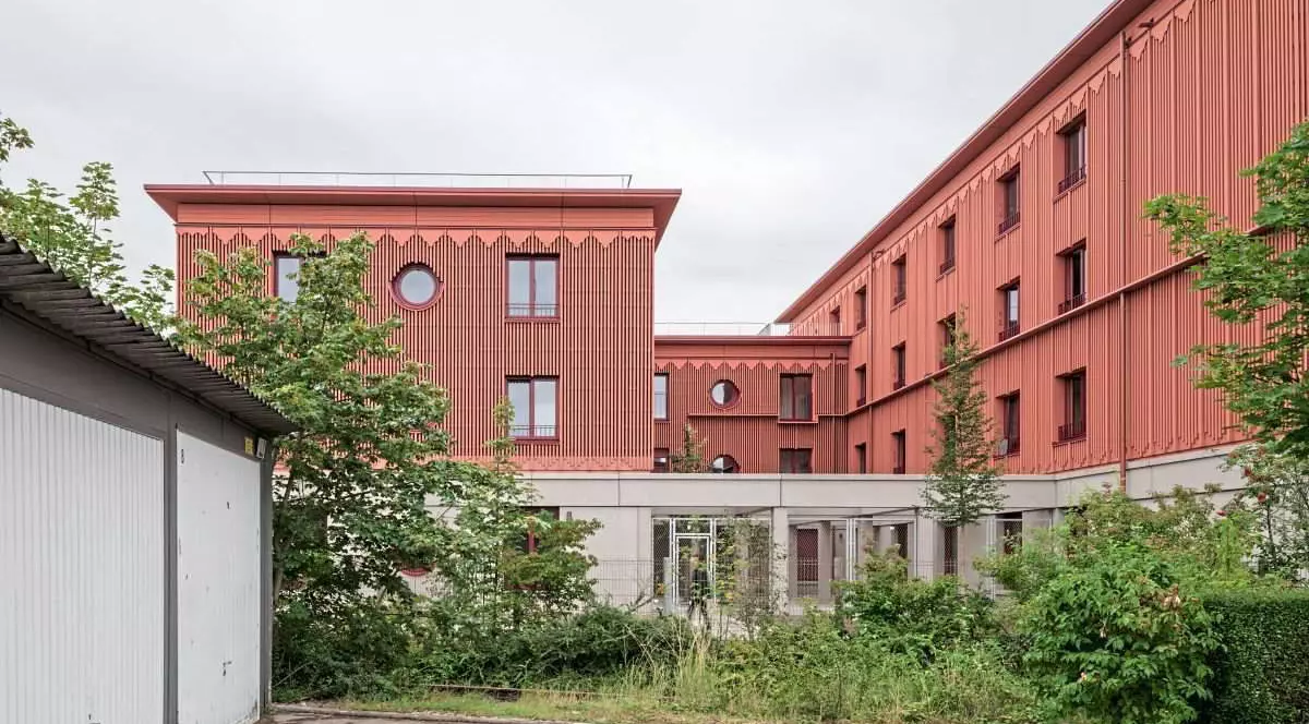 Un adăpost roșu cărămiziu pentru persoane fără casă, în Munchen, situat la periferie și într-o zonă industrială. În fața clădirii sunt câțiva pomi verzi