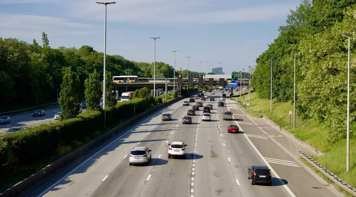 Autostradă aglomerată cu mai multe benzi pe sens. Traficul este fluid, iar printre vehiculele aflate în mișcare se observă autoturisme, un autobuz și un camion. În fundal se vede o pasarelă rutieră.
