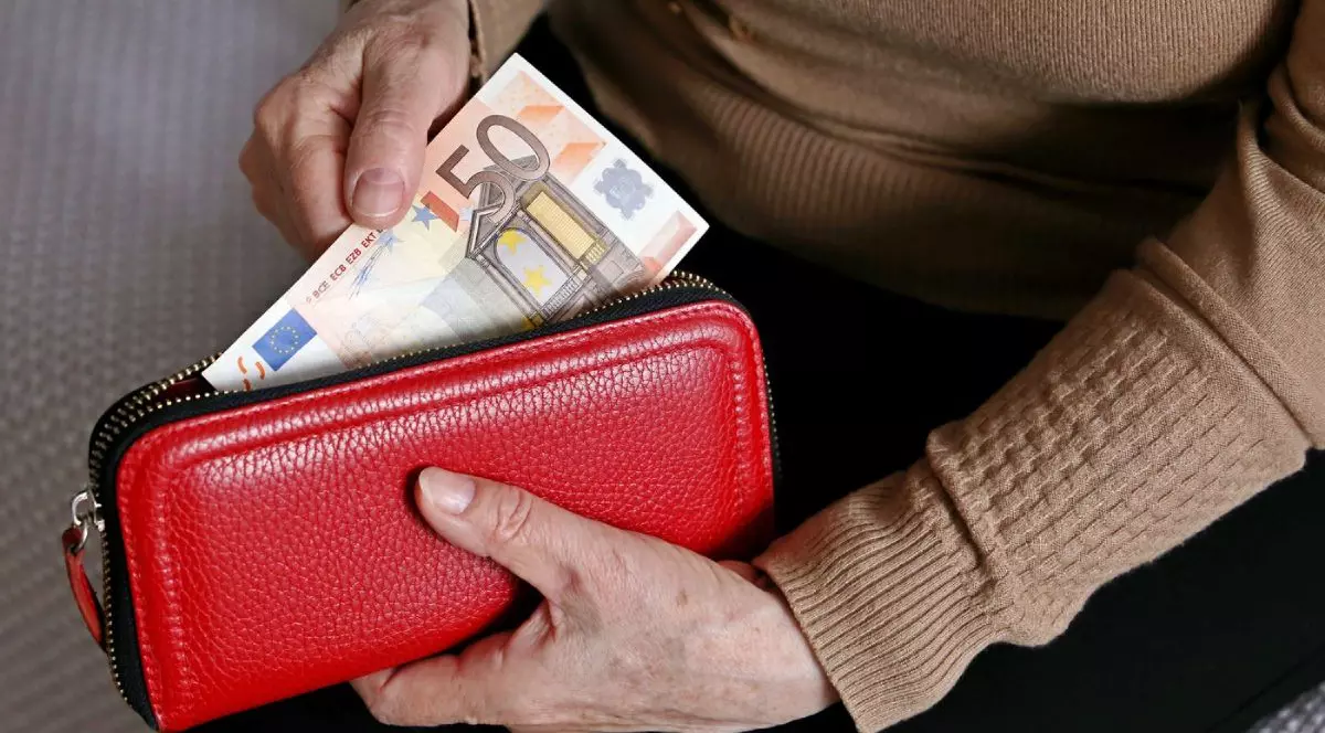 O femeie îmbrăcată cu un pulover de culoare maro scoate o bancnotă de 50 de euro dintr-un portofel de culoare roșie