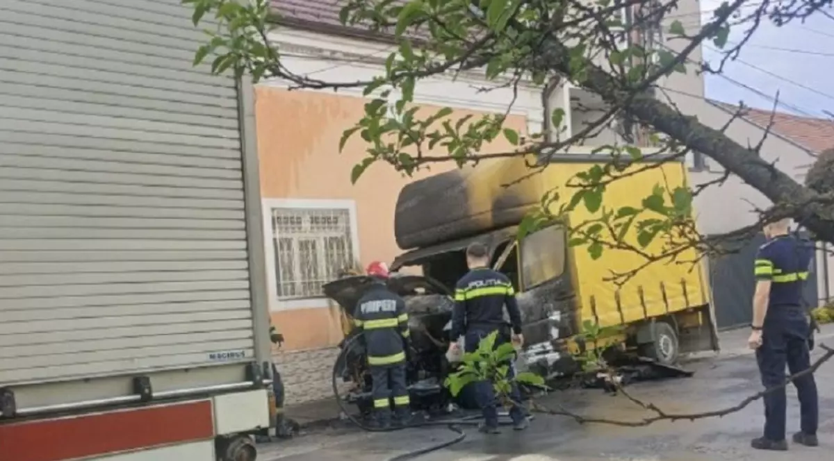 În imagine este ilustrată o camionetă galbenă, care a ars. De asemenea, se observă și pompieri și polițiști în apropierea acesteia