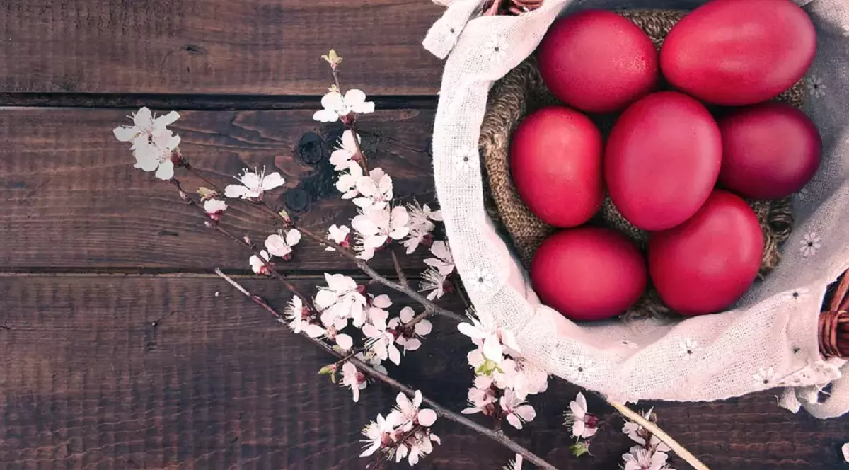 Coș alb cu ouă roșii de Paște, pe o masă de lemn maronie presărată cu flori albe