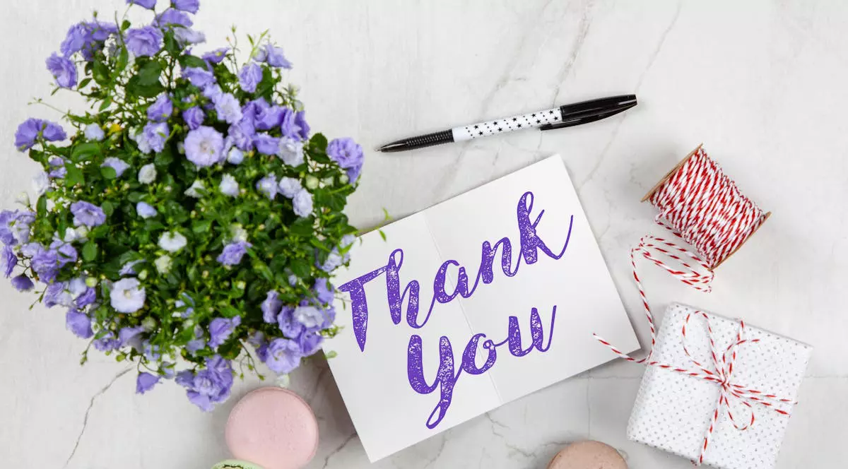 Cadou nou nascut: poza cu un card pe care scrie Thank you cu mov, avand in stanga un buchet de flori, in dreapta un cadou si cateva macarons aruncate in partea de jos
