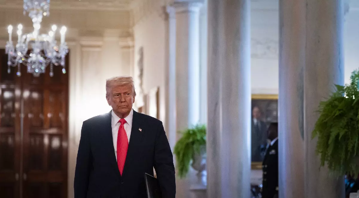 Donald Trump, îmbrăcat într-un costum albastru închis, cămașă albă și cravată roșie, mergând pe un hol. În dreapta lui sunt niste ferestre cu perdele albe, în stânga sus este un candelabru.