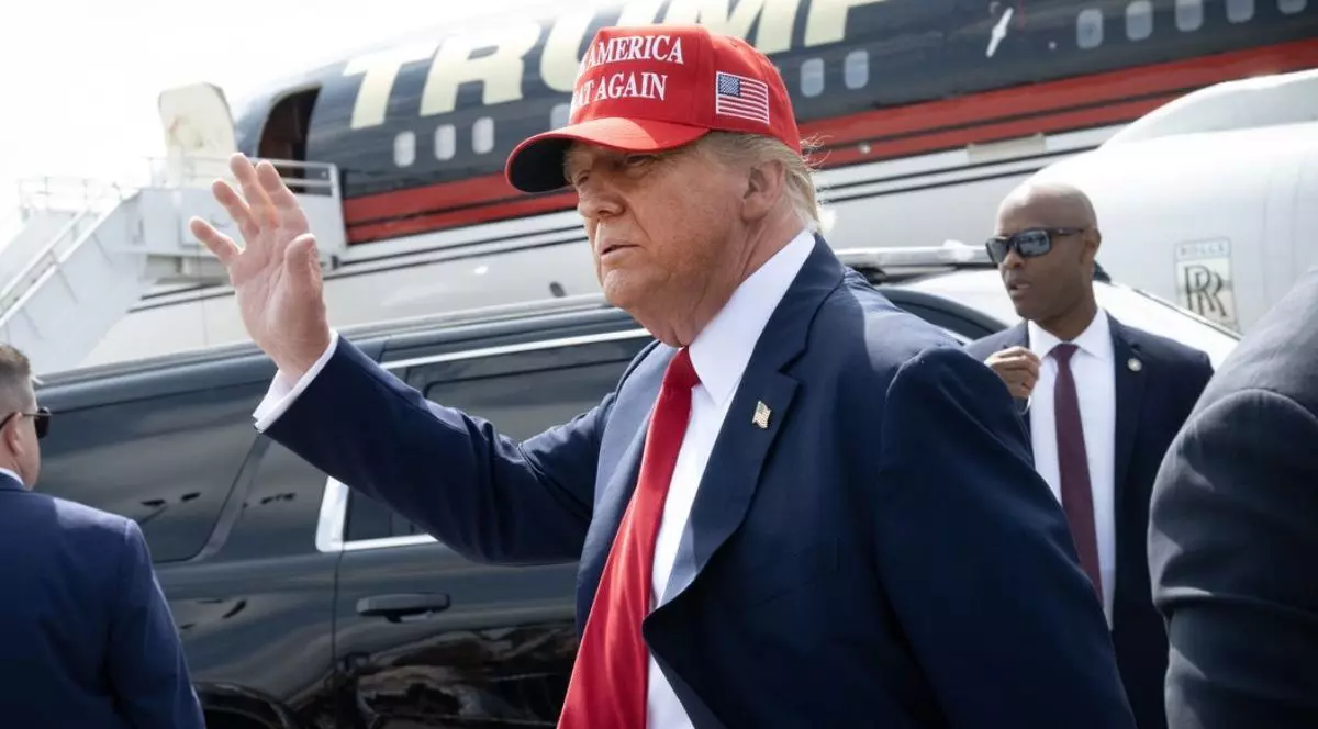 Donald Trump, pe aeropost, lângă un avion, în costum negru cu o cravată roșie si cu o paparie tot rosie, cu mâna dreaptă ridicată