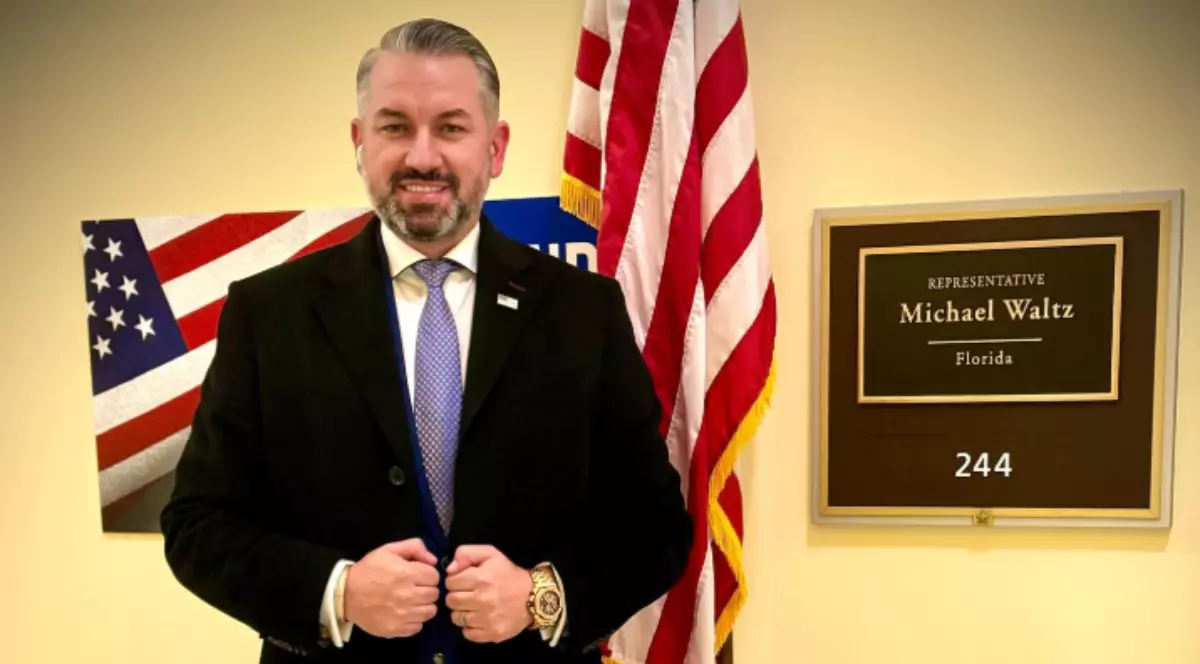 În imagine este ilustrat un bărbat care zâmbește. Acesta este îmbrăcat într-un palton negru, poartă o cămașă albă și are o cravată albastră.