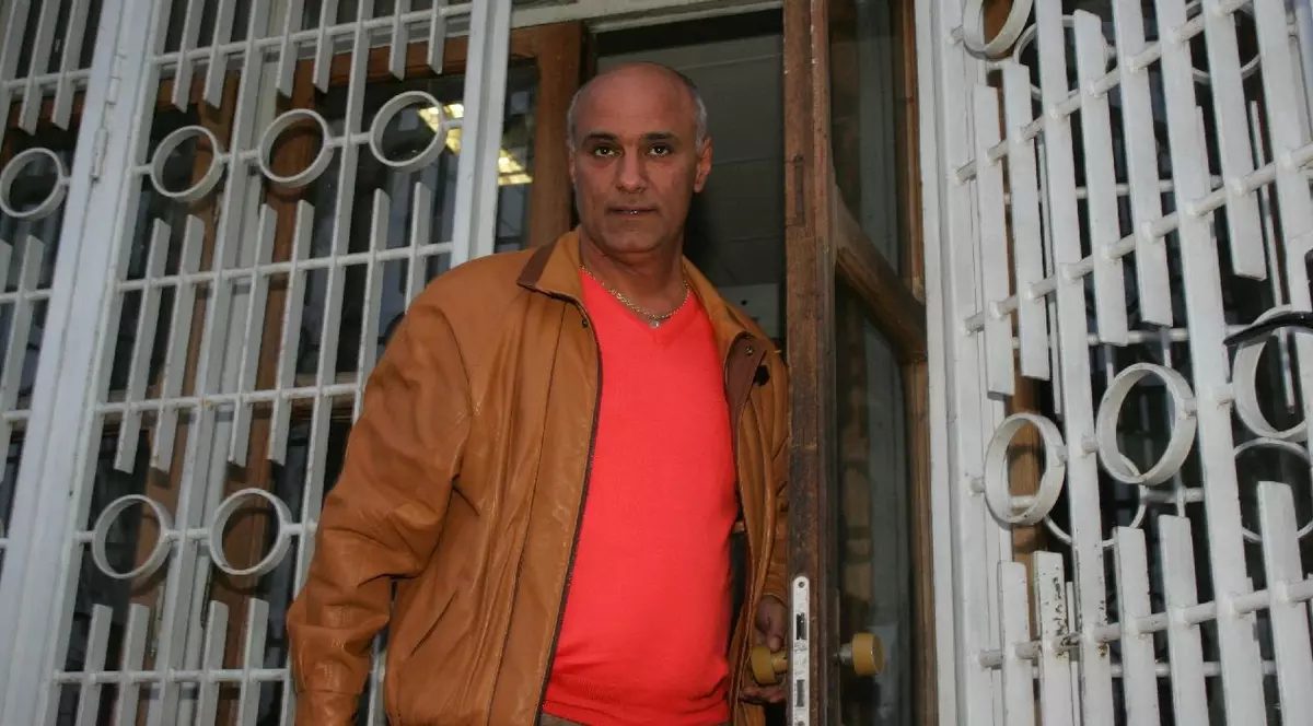 Un bărbat cu geacă maro și tricou portocaliu iese pe o poartă din fier