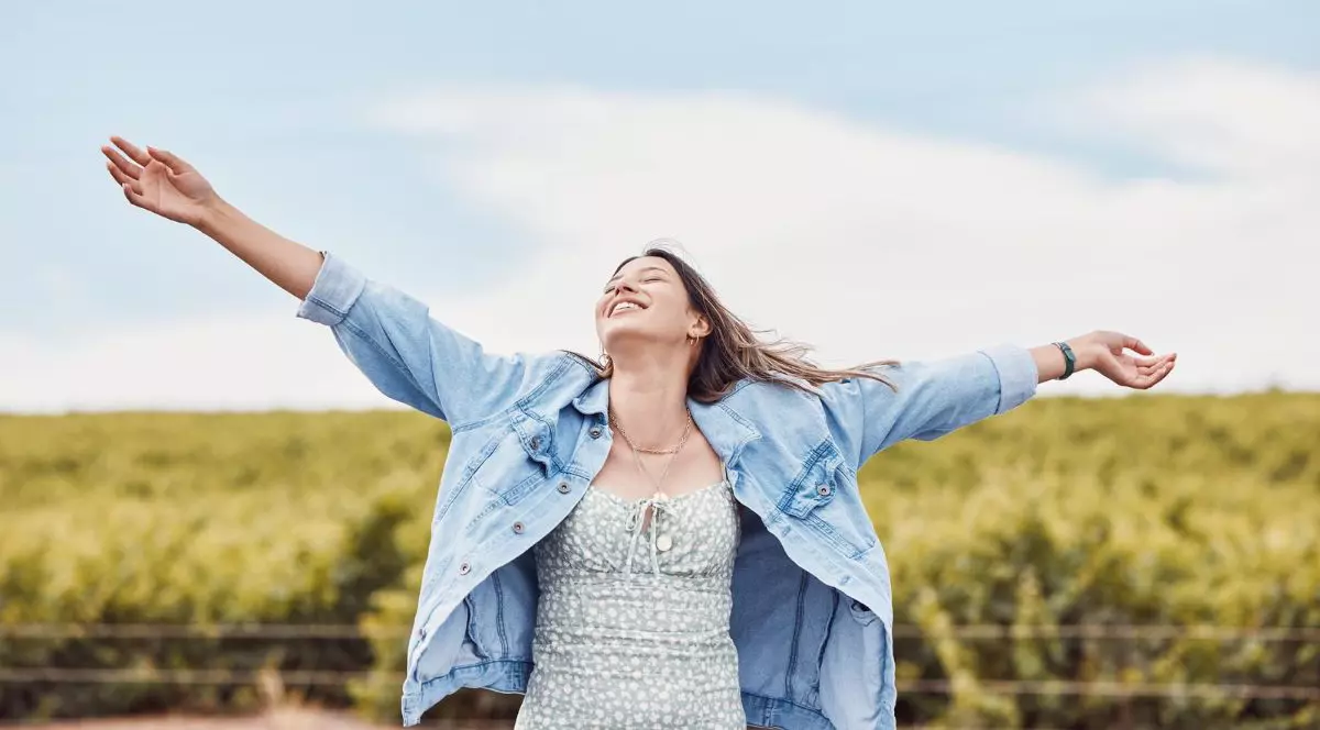 femeie îmbrăcată într-o rochie bleu și o geacă de blugi largă, cu mâinile deschise, în timp de zâmbește, aflată pe un câmp verde cu flori
