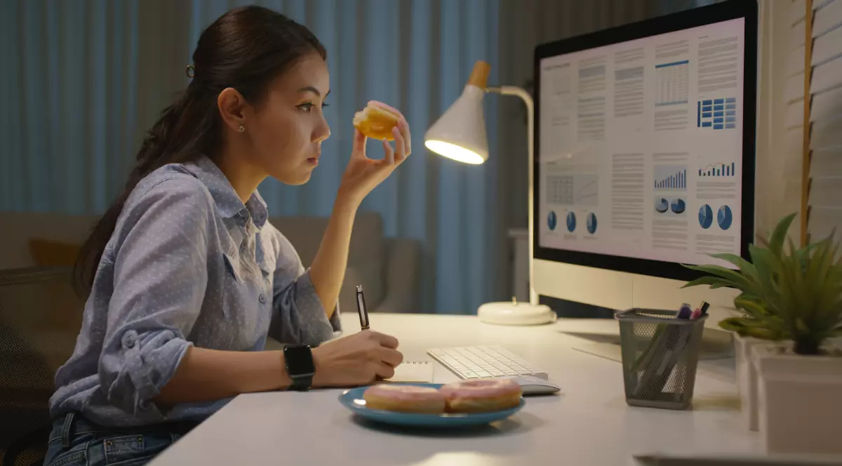 O femeie îmbrăcată într-o cămașă albastră, lucrează la un birou pe timp de noapte, în timp ce mănâncă o gogoașă.