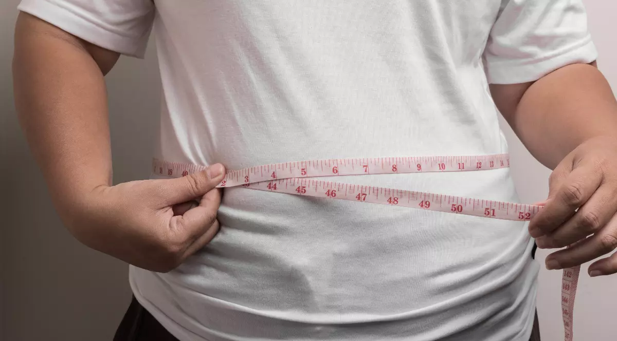 În imagine este ilustrată o femeie îmbrăcată în tricou alb, care își măsoară talia