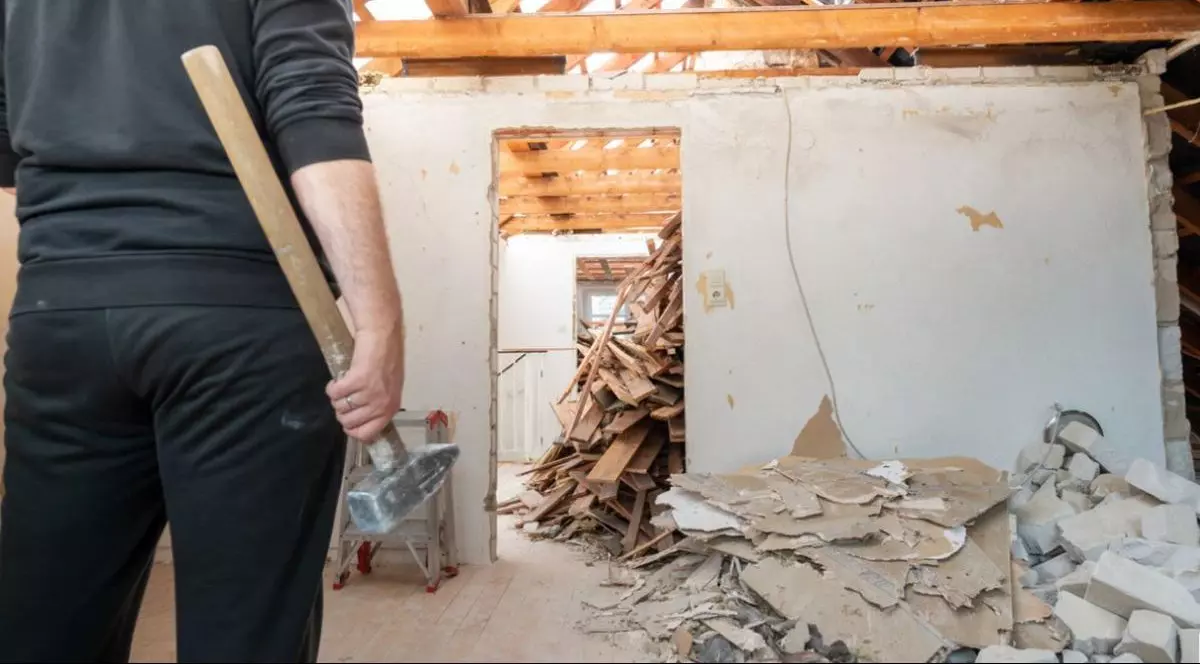 Un bărbat fotografiat cu spatele, îmbrăcat în negru, și cu un ciocan în mână este într-o casă în care e demolată o parte din ea