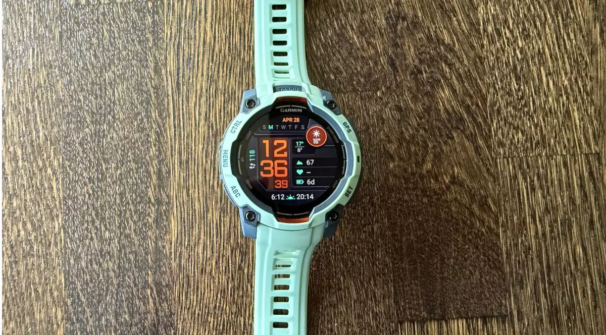 Ceas Garmin Instinct 3 AMOLED, de culoare turcoaz, așezat pe un fundal de lemn.