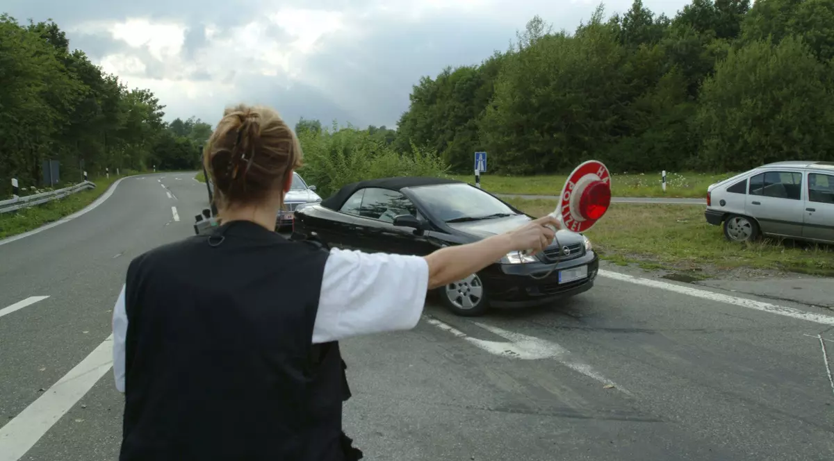 Poză cu o polițistă care trage pe dreapta o mașină neagră, pe o șosea din Germania. Ține în mâna dreaptă o paletă roșie și este fotografiată din spate