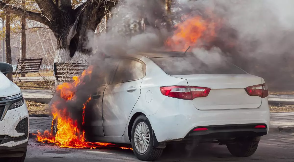 Masina de culoare alba in flacari, langa ea este un copac si o alta masina