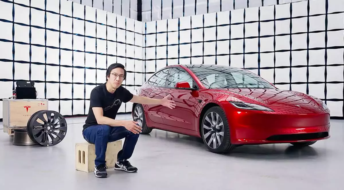 un barbat care sta asezat pe un cub, imbracat in negru, in spatele lui este o masina tesla rosie