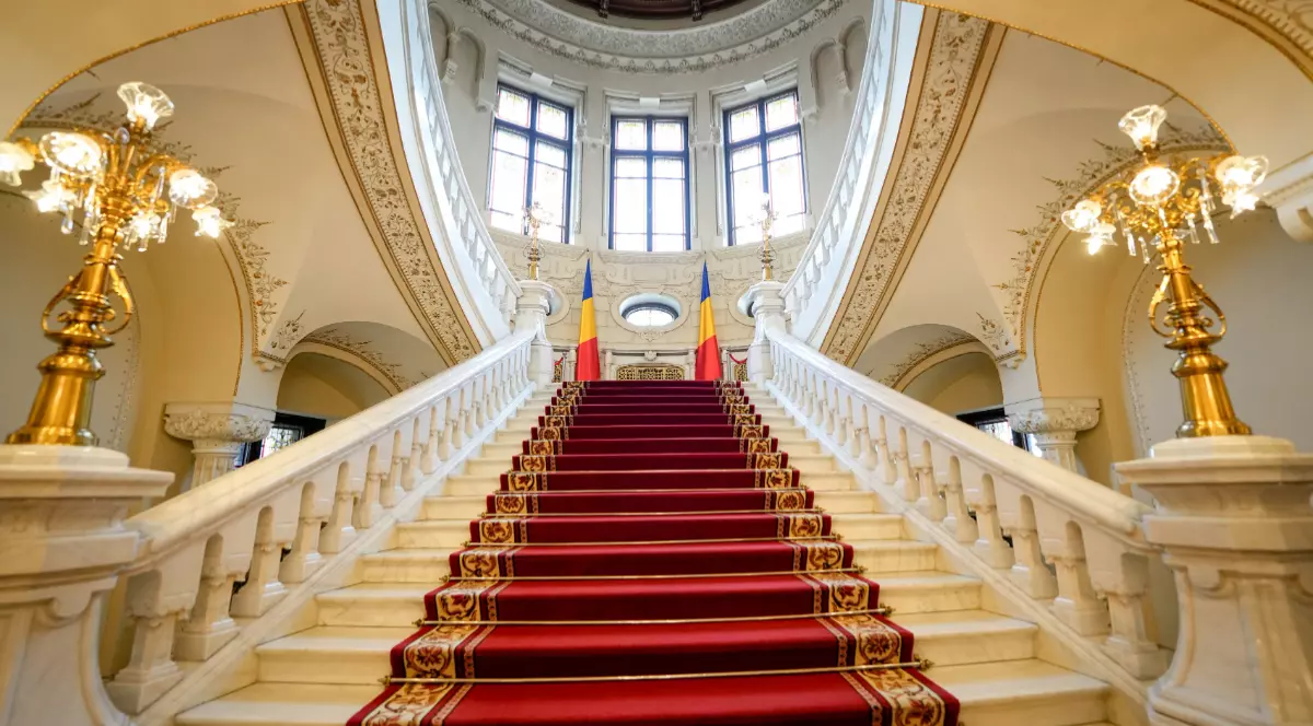 Imaginea prezintă o scară din marmură albă, acoperită cu un covor roșu, care urcă simetric spre un etaj superior luminat de un ansamblu de ferestre mari