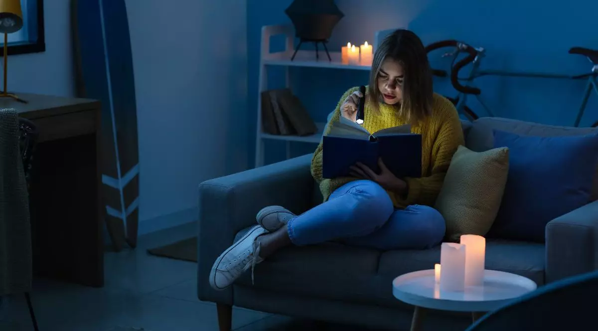 O tânără stă într-un fotoliu și citește o carte la lumina lumânărilor și ținând în mână o lanternă, în timpul unei pene de curent