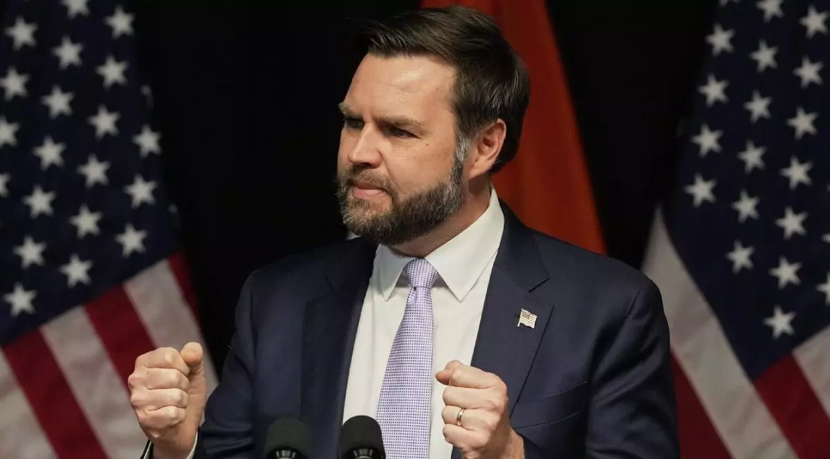 JD Vance in timp ce vorbește, mâinile ridicate și pumnii strânși, în costum închis la culoare și cravata gri deschis, cu steagul SUA în spate