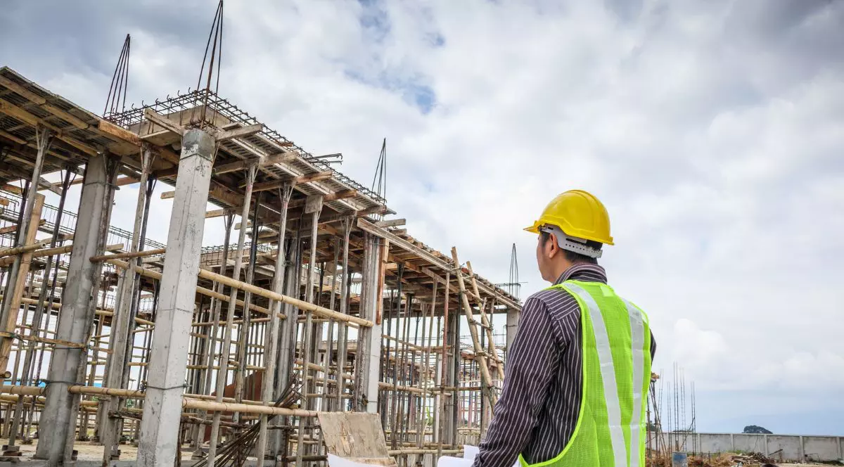 clădire impobiliară în construcții, privită de un inginer îmbrăcat în vestă reflectorizantă cu o cască de constructor pe cap