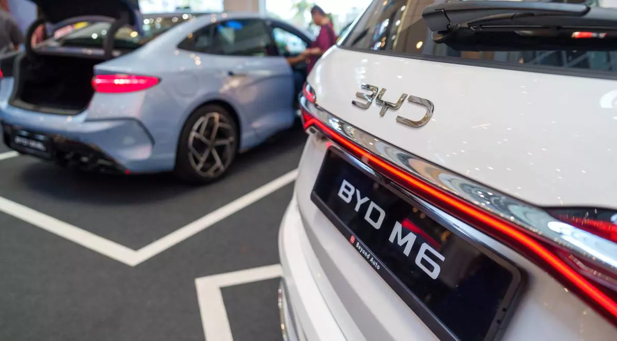 Două mașini electrice expuse într-un showroom sau la un eveniment auto. În prim-plan partea din spate a unei mașini albe marca BYD, modelul M6, evidențiat de logo-ul mare „BYD” și stopurile LED. În fundal este oa ltă mașină de culoare albă.