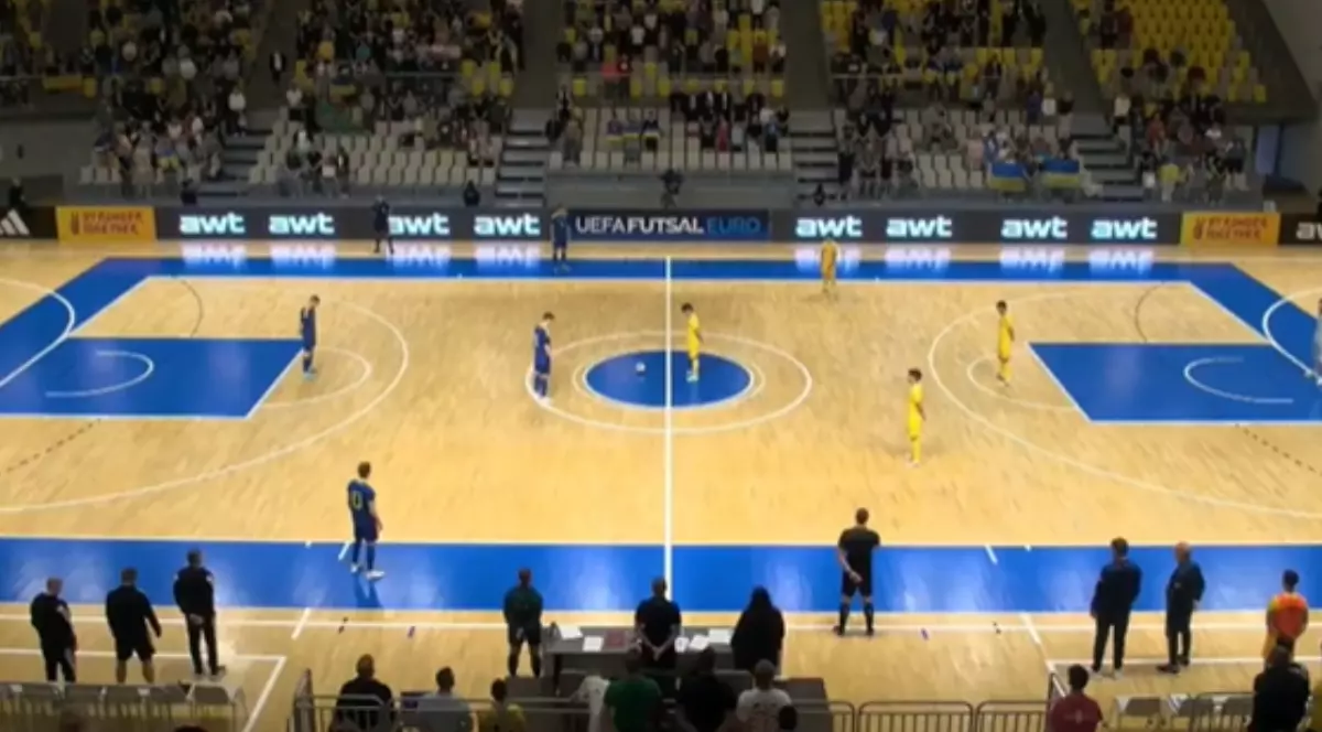 Imaginea surprinde un teren de futsal. În partea stângă sunt jucători îmbrăcați în albastru, iar în partea dreaptă sunt jucători în echipament galben. În fundal se observă tribuna cu câțiva oameni așezați pe scaune.