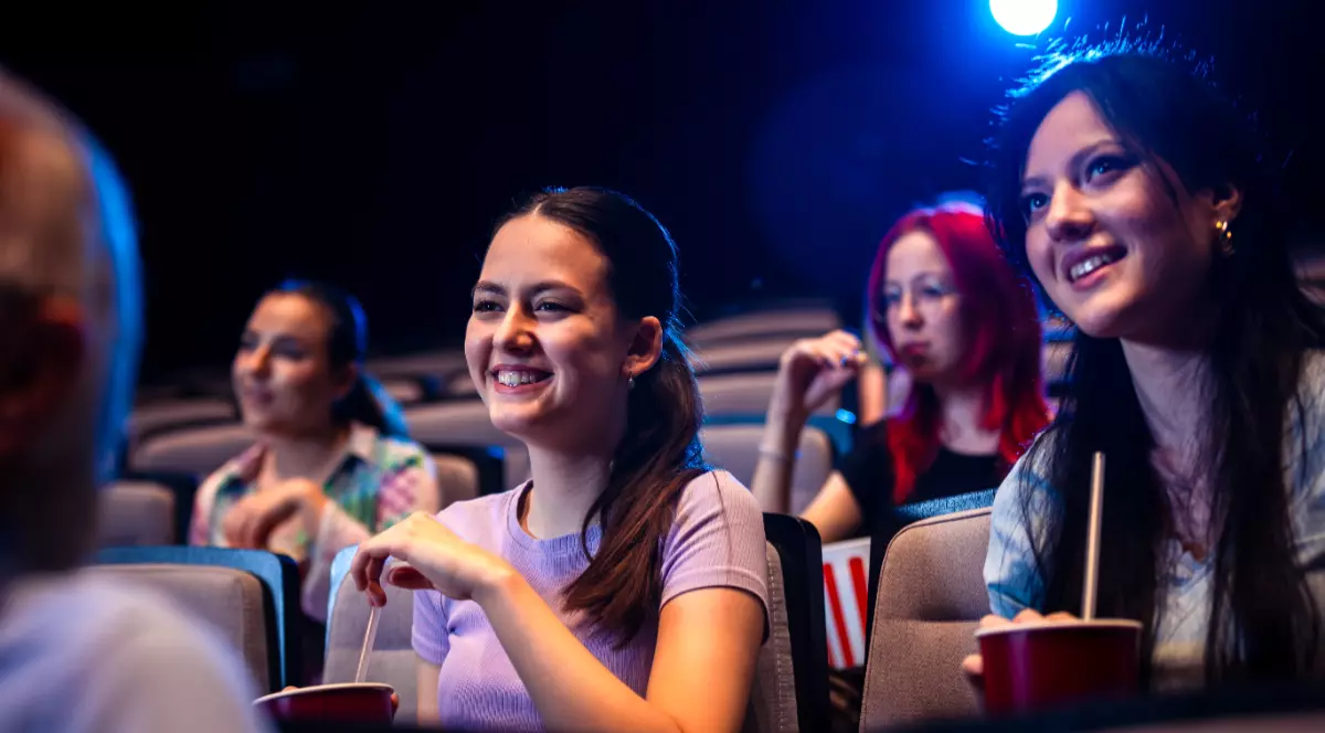 În imagine sunt ilustrate fete care se află pe scaune într-o sală de spectacol. Acestea zâmbesc și țin în mână pahare cu paie.
