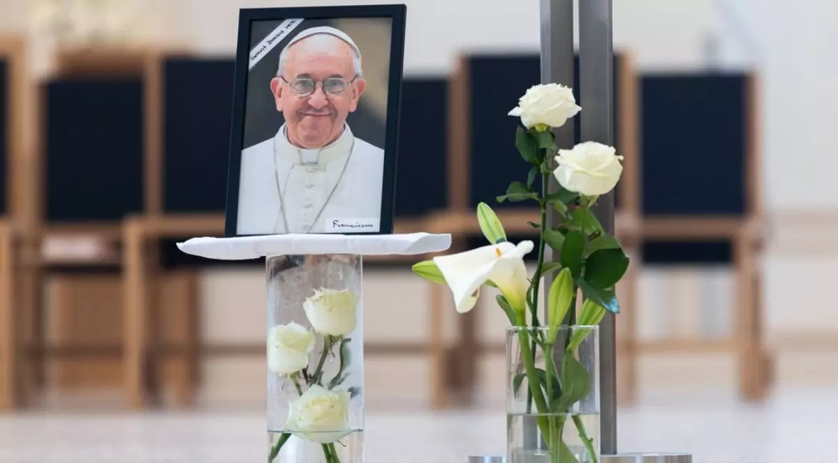 Papa Francisc a murit, imagine cu o fotografie cu Papa pe un pahar cu flori alve, alaturi un alt pagar cu trandafiri albi iar in spate niste scaune negre
