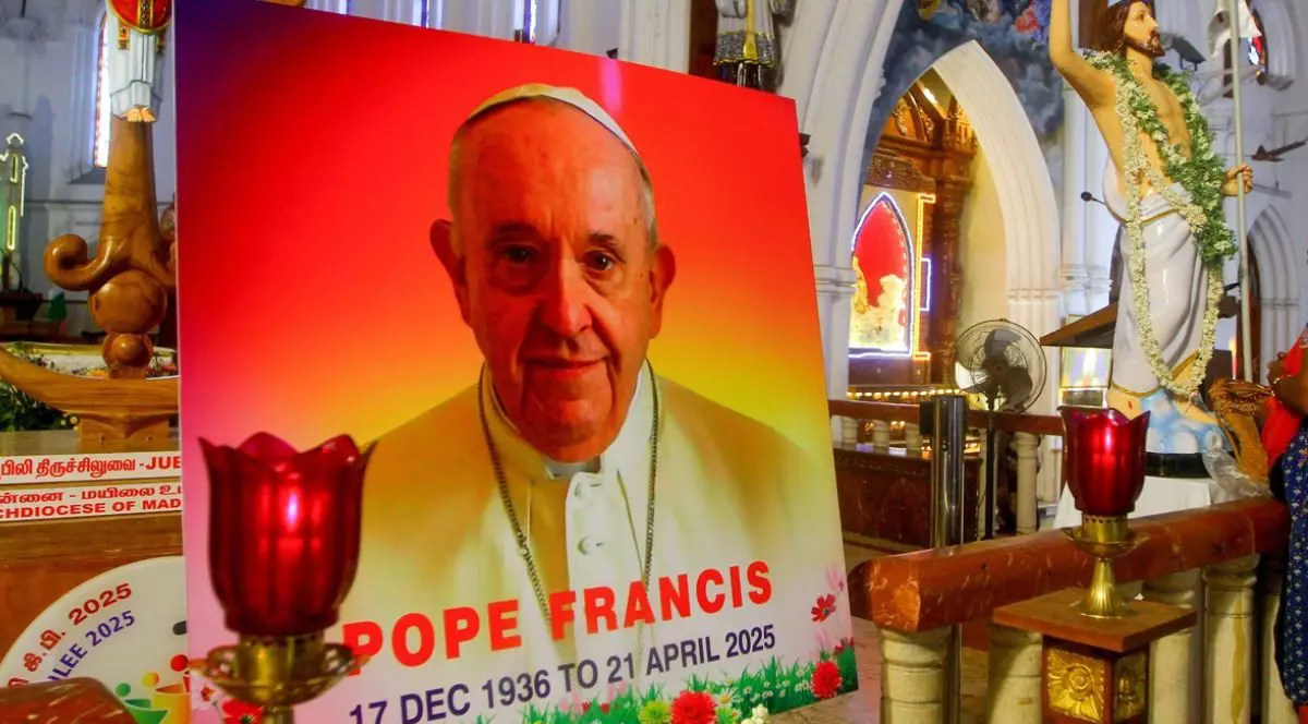 Un portret al Papei Francisc, cu scris pe el și o candelă aprinsă, o statuie a lui Isus Cristos în spate, într-o biserică