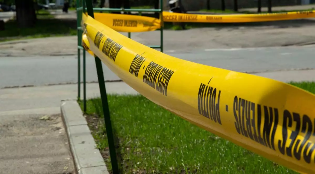 În prim-plan se vede o bandă galbenă, cu inscripția „Poliție, acces interzis”, cu care este împrejmuită o zonă verde de pe o stradă.