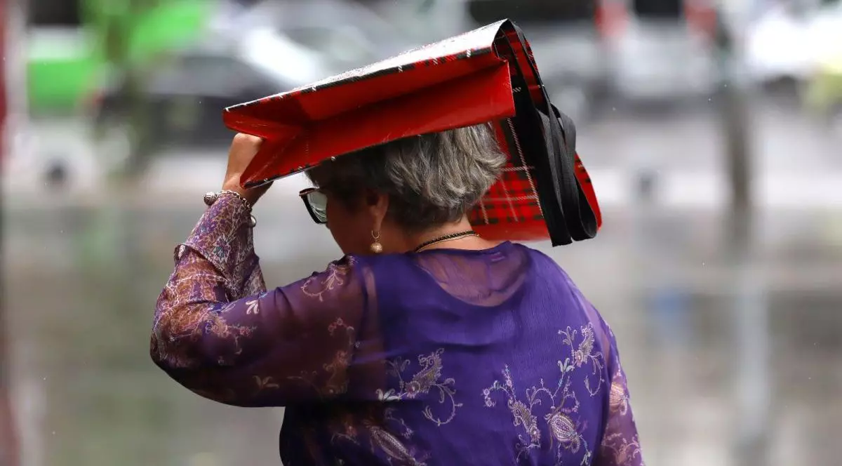 O femeie, fotografiată cu spatele, merge pe stradă și a prins-o ploaia, astfel că își ține cu mâna o sacoșă cu care își acoperă capul.