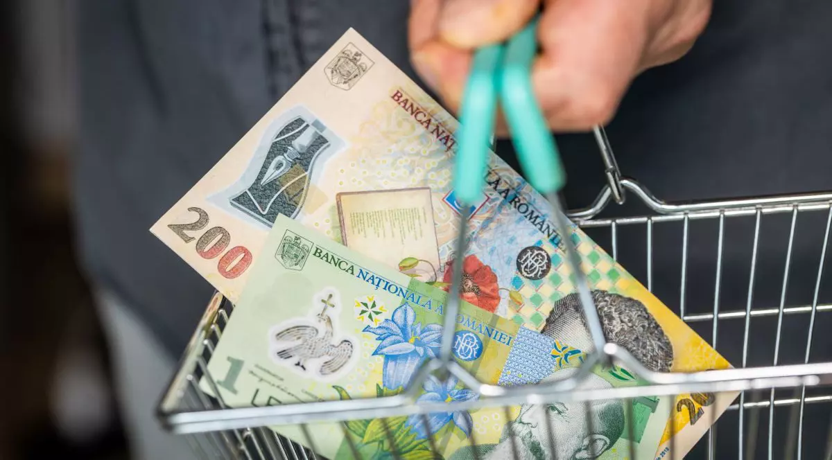 Un bărbat ține în mână un coș de cumpărături de mărime foarte mică, în care se vede o bancnotă de 200 de lei și una de un leu