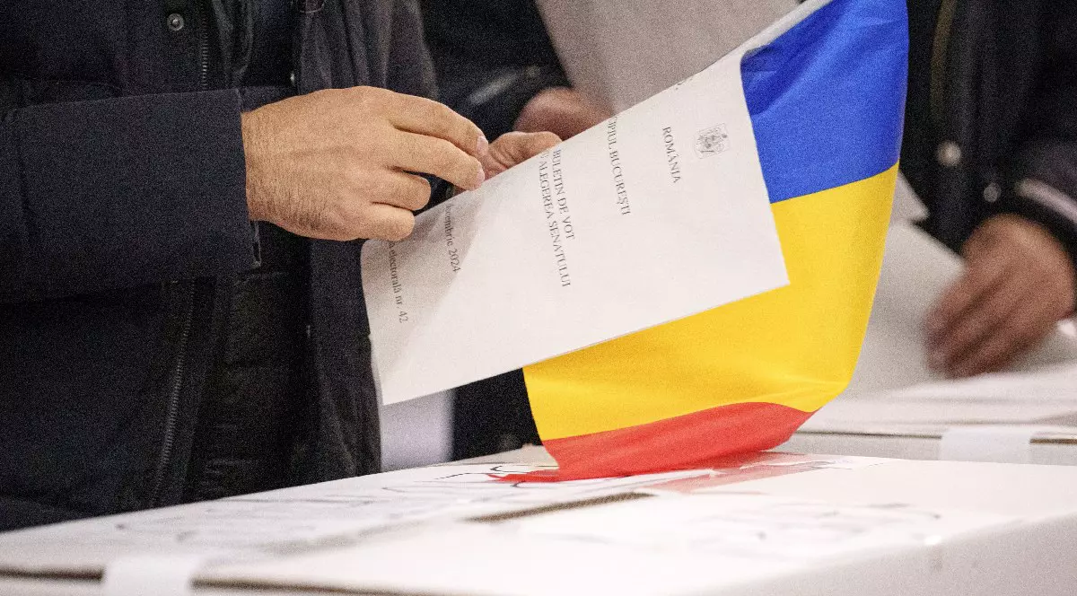 Un român are buletinul de vot, ștampila și drapelul țării într-o mână și se pregătește să bage în urnă hârtia pe care a votat