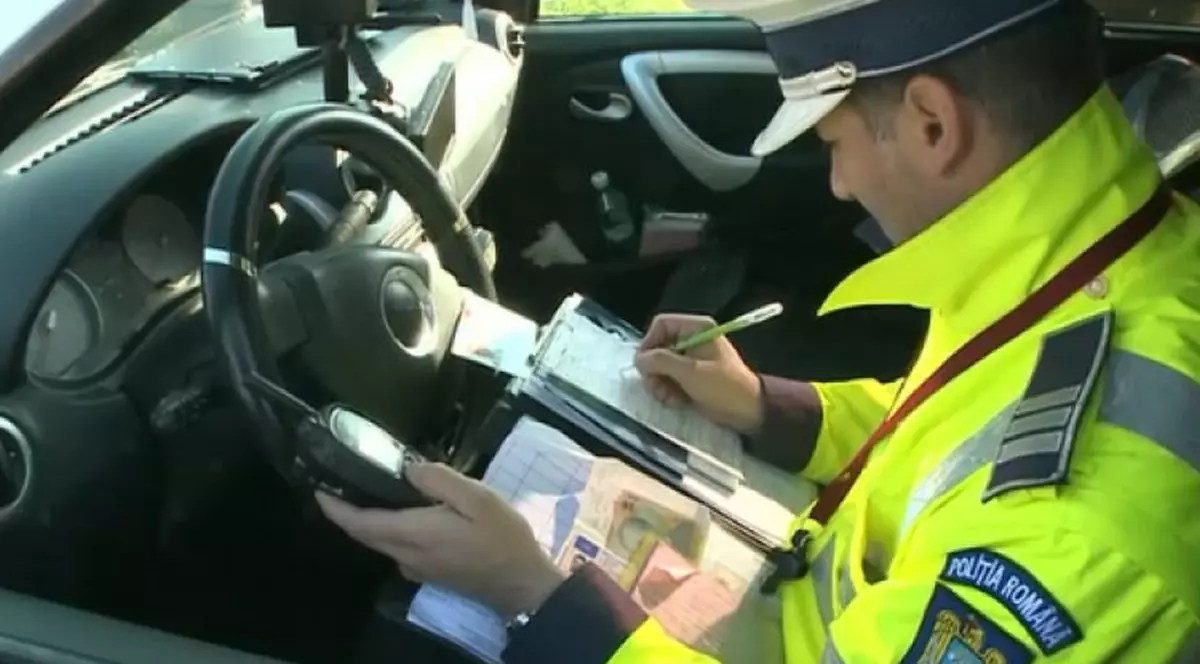Un polițist cu uniformă este în mașină și scrie o amendă pentru un șofer, iar lângă el are niște bancnote