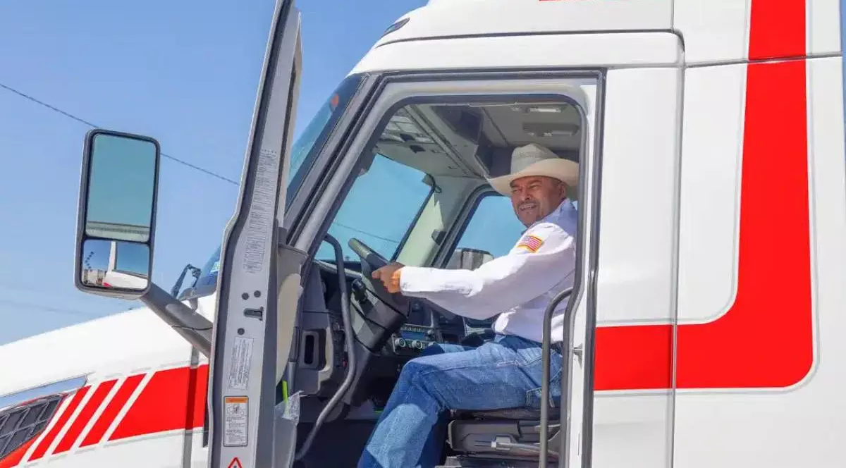 Șofer de camion american în cabina vehiculului. Bărbatul poartă o pălărie tip cowboy, o cămașă albă cu un mic drapel american pe mânecă și blugi albaștri.