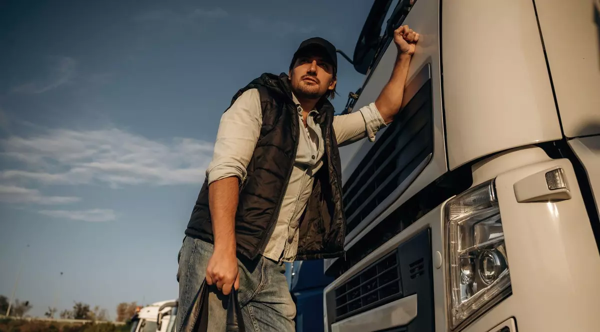 Poză cu un șofer care se sprijină cu o mână de un camion. Are în mână o geantă