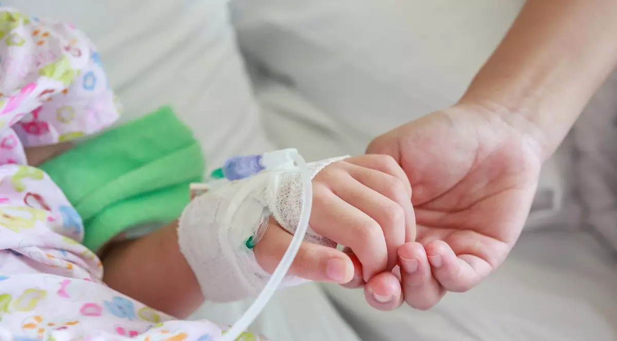 mama care ține de mână un copil în spital, cu o branulă în mână