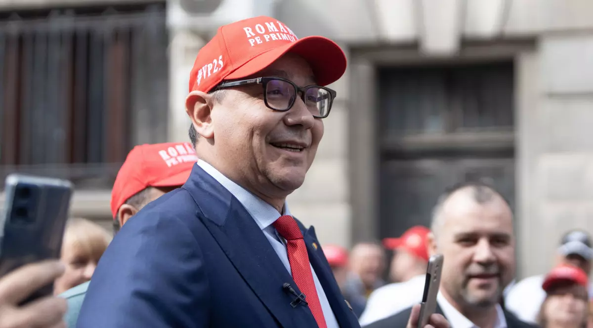 Victor Ponta îmbrăcat într-un sacou albastru, cu cămașă albă, cravată roșie și o șapcă roșie pe cap. În spatele său sunt mai multe persoane
