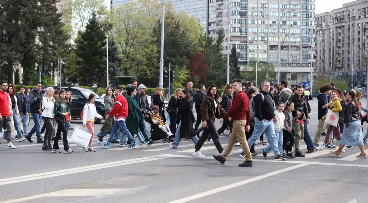 Mulți oameni traverseazaă strada pe la trecerea de pietoni, iar în jur se văd blocuri și copaci cu frunze verzi, București, 13 aprilie 2025