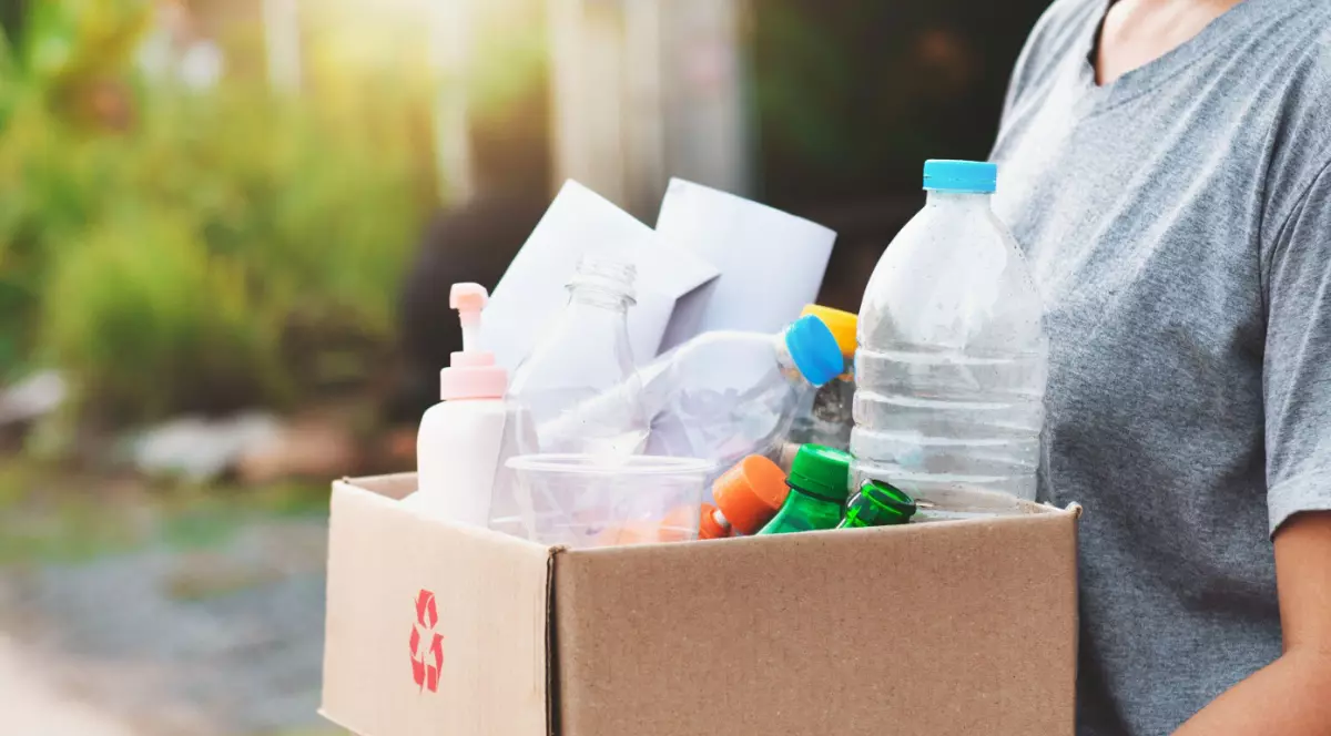 Barbat care care o cutie de carton in care se afla diferite sticle PET de reciclat