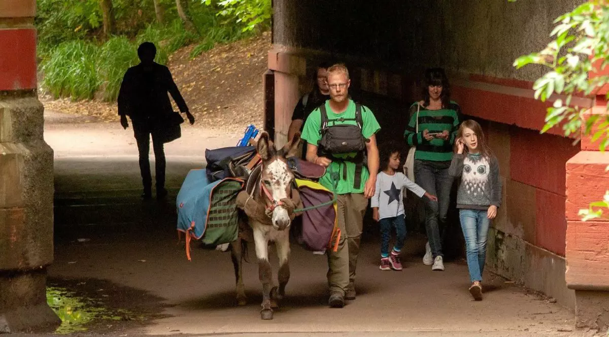 Un bărbat pe nume Adam, în tricou verde, alături de măgărusul său, Marin, incarcat cu bagaje, merg pe jos într-o calatorie