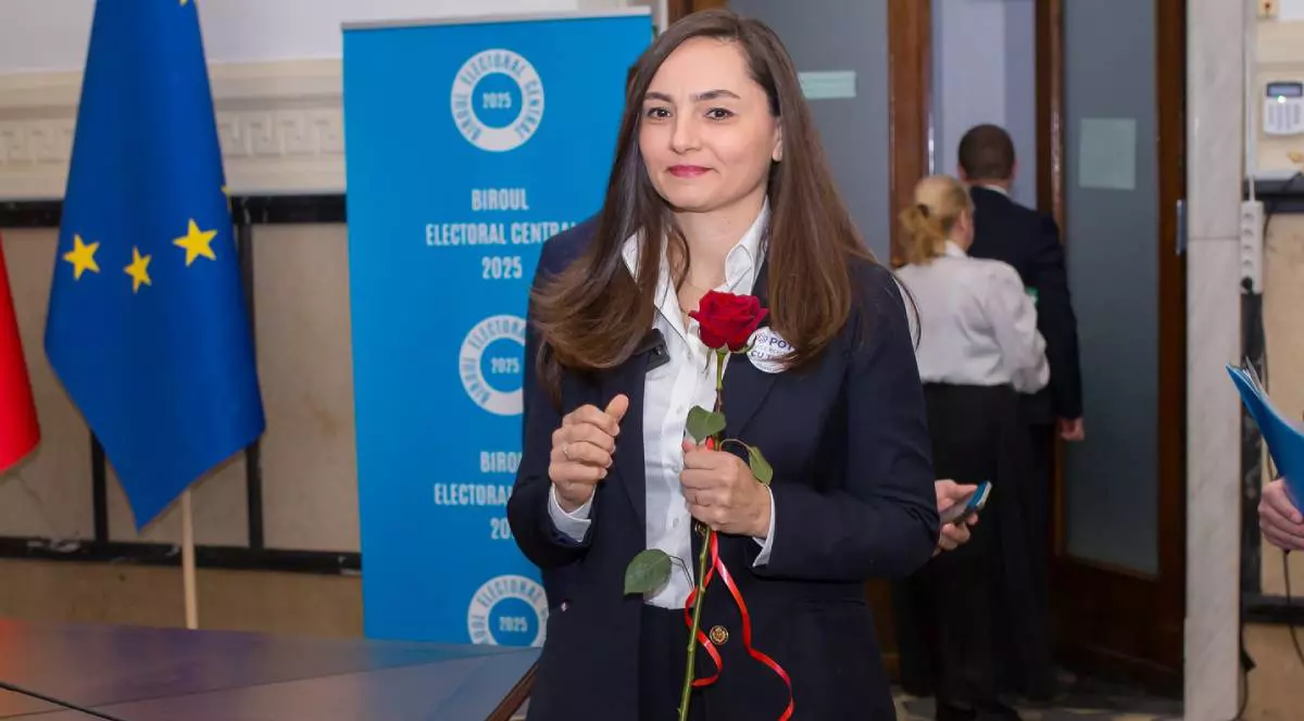 Anamaria Gavrilă îmbrăcată într-un costum negru și cămașă albă, în timpul ce ține un trandafir roșu în mână, surprinsă veselă la Biroul Electoral Central.