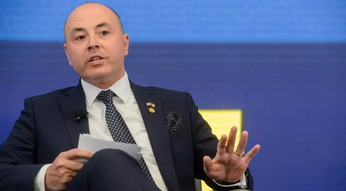 Ambasadorul României în SUA, George Muraru, stă pe un fotoliu, ține niște foi în mână și gesticulează cu cealaltă mână, iar în spate are un fundal albastru