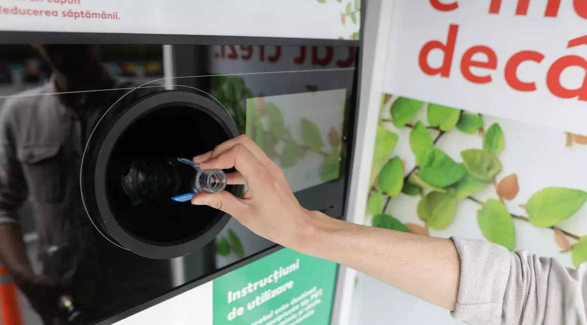 O persoană care introduce o sticlă de suc din sticlă, fără capac, într-un aparat de reciclare al sticlelor.