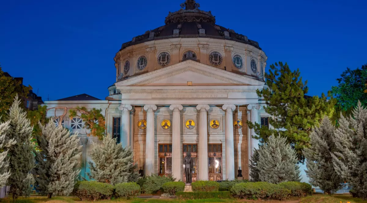 În imagine este ilustrat Ateneul Român. Clădirea este fotografiată frontal, seara.