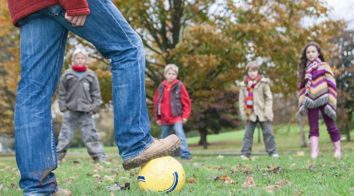 Imagine cu picioarele unui bărbat care tine o minge sub piciorul drept, in fundal patru copiii care așteapta