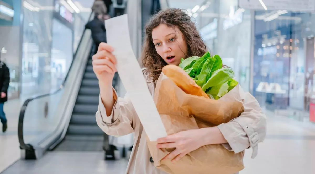 O femeie se uită foarte mirată la un bon de cumpărături într-un supermarket dintr-un mall și are în brațe un pachet cu pâine și salată, iar în fundal se văd scări rulante