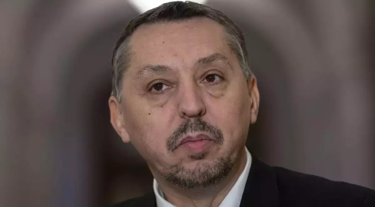 Imaginea arată un bărbat de vârstă mijlocie, îmbrăcat formal, purtând un sacou negru și o cămașă albă.