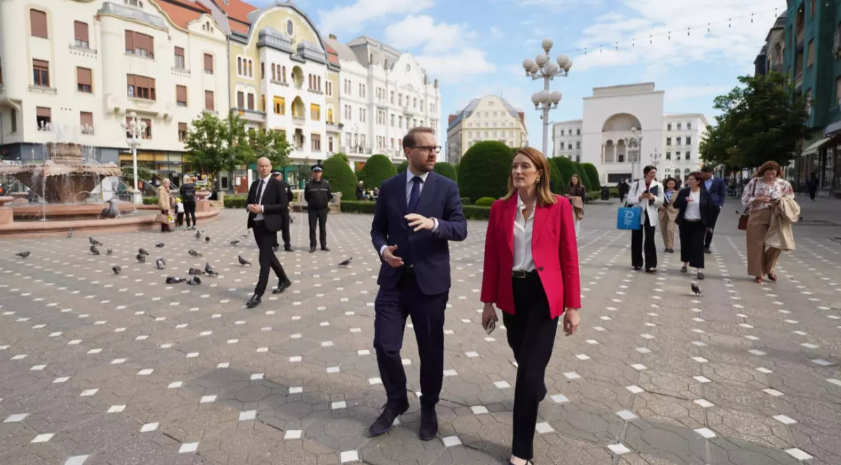În poză, Roberta Metsola și primarul Dominic Fritz se plimbă prin centrul Timișoarei și vorbesc. Sunt înconjurați de câțiva oameni, iar în fundal se văd clădiri și o fântână.