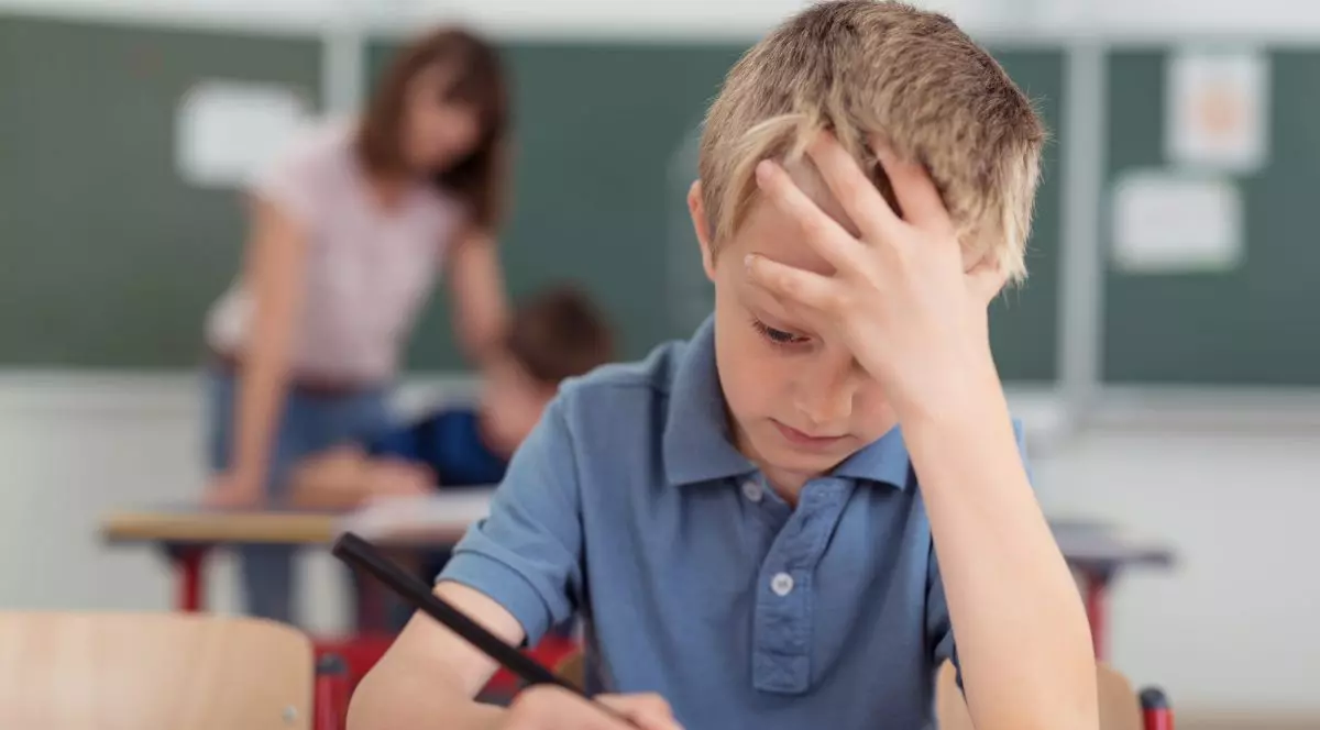 Elev de vârstă mică care stă în bancă, cu mâna în cap și se concentreaza la un test.