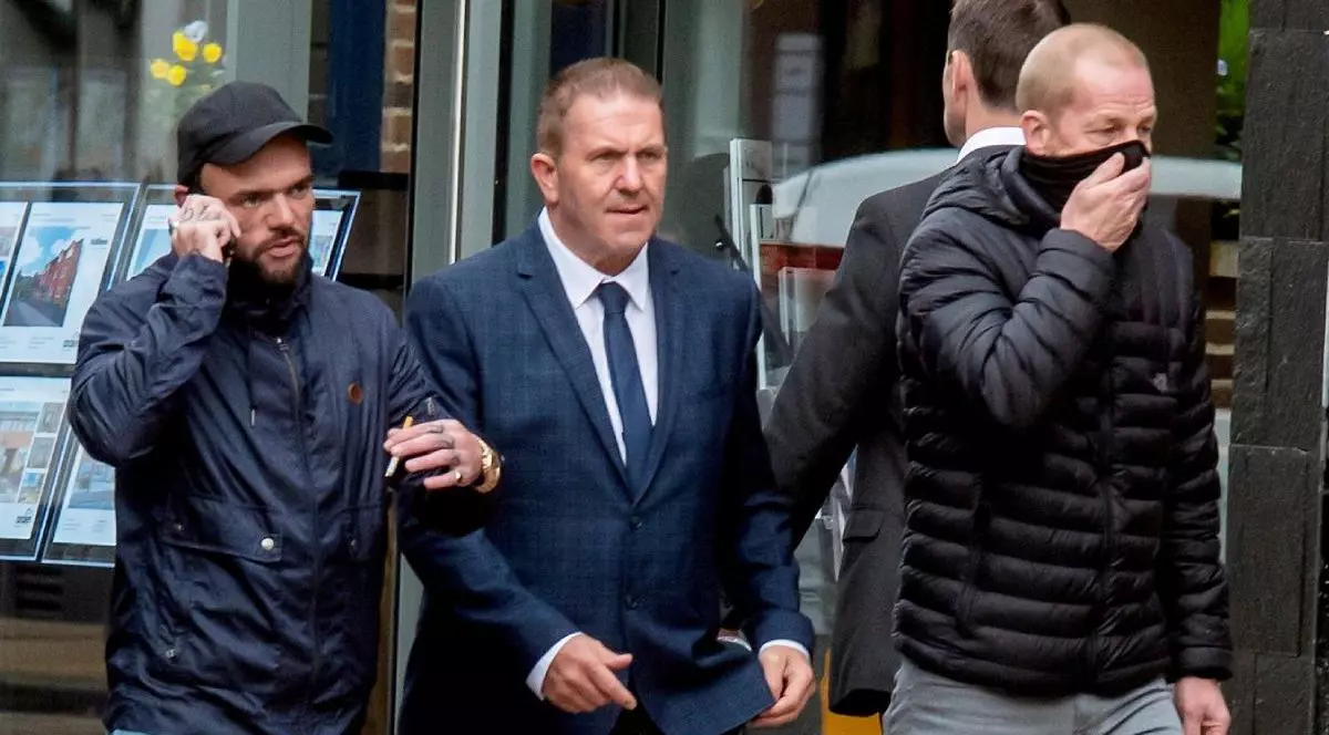 Trei barbati merg oe strada, toti îmbrăcați in haine inchise la culoare, primul din stanga cu sapca si vorbeste la telefon, cel din mijloc in costum si cravata, cel din dreapta cu geaca si fata acoperita , in spatele lor o vitrina cu fotografii - tablouri